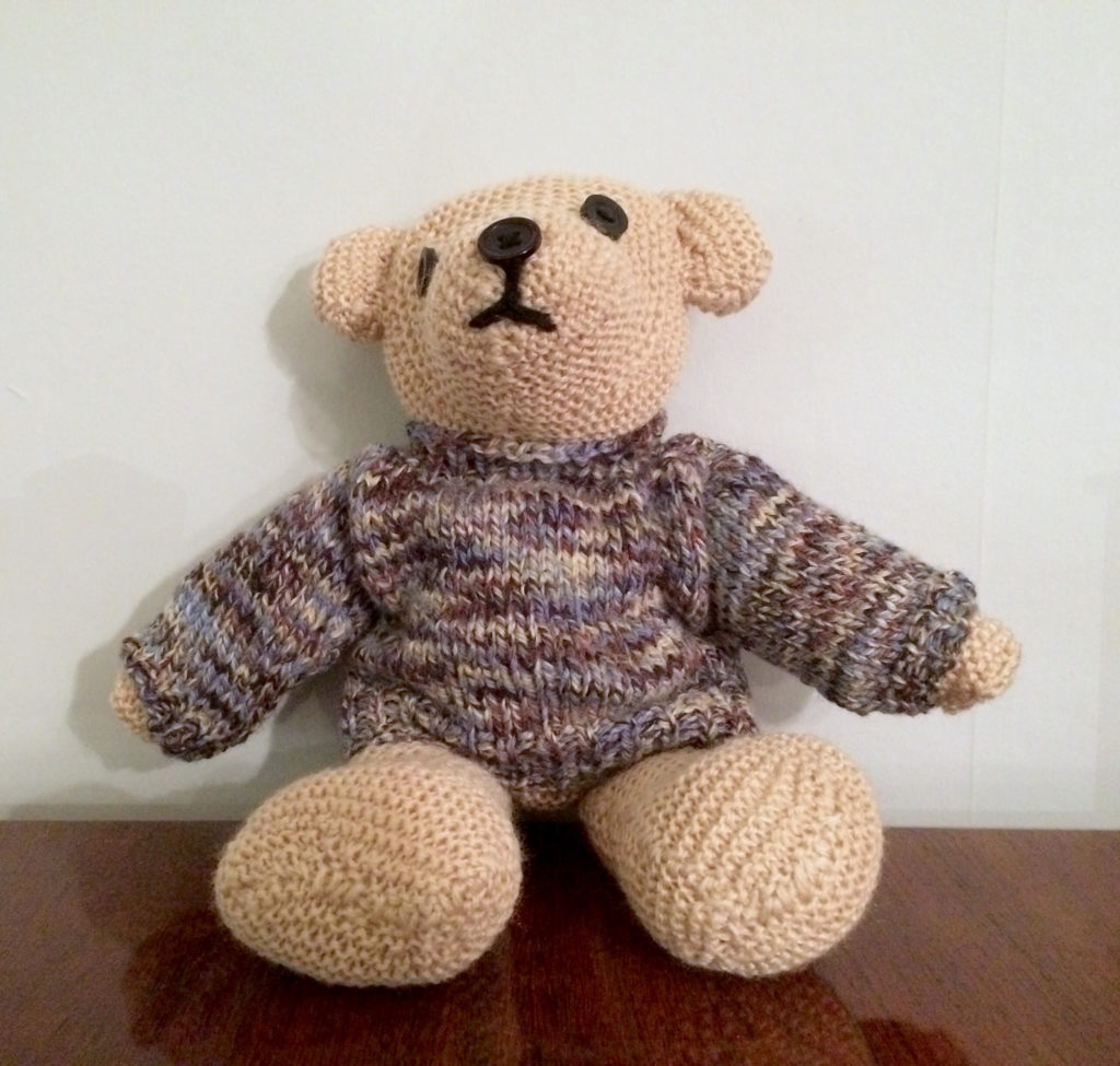 Knitted teddybear with a slightly wonky face wearing a multicoloured knitted jumper