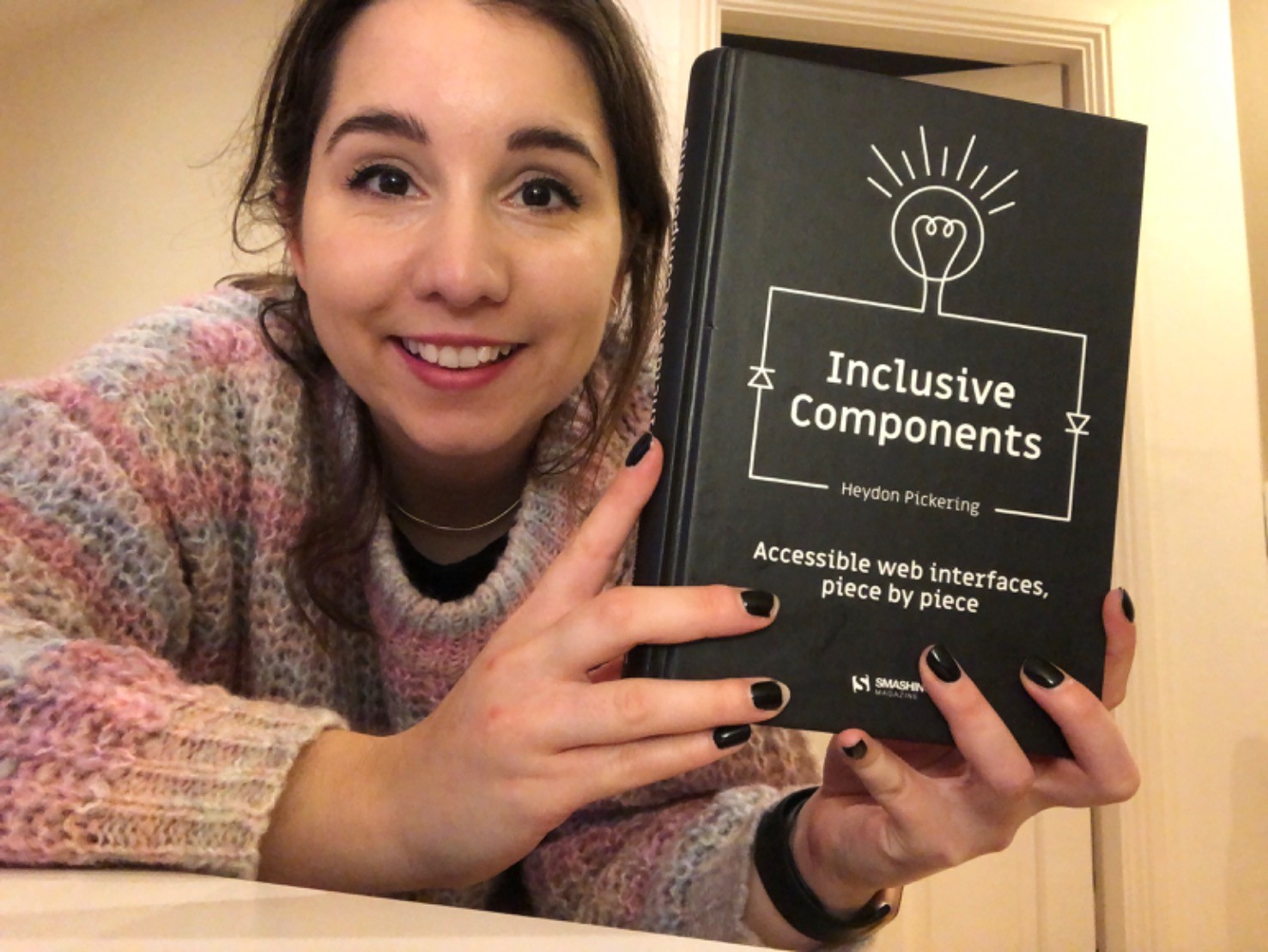 Me clutching a hardback copy of Heydon Pickering’s Inclusive Components book. The book is black and my nails are painted black.