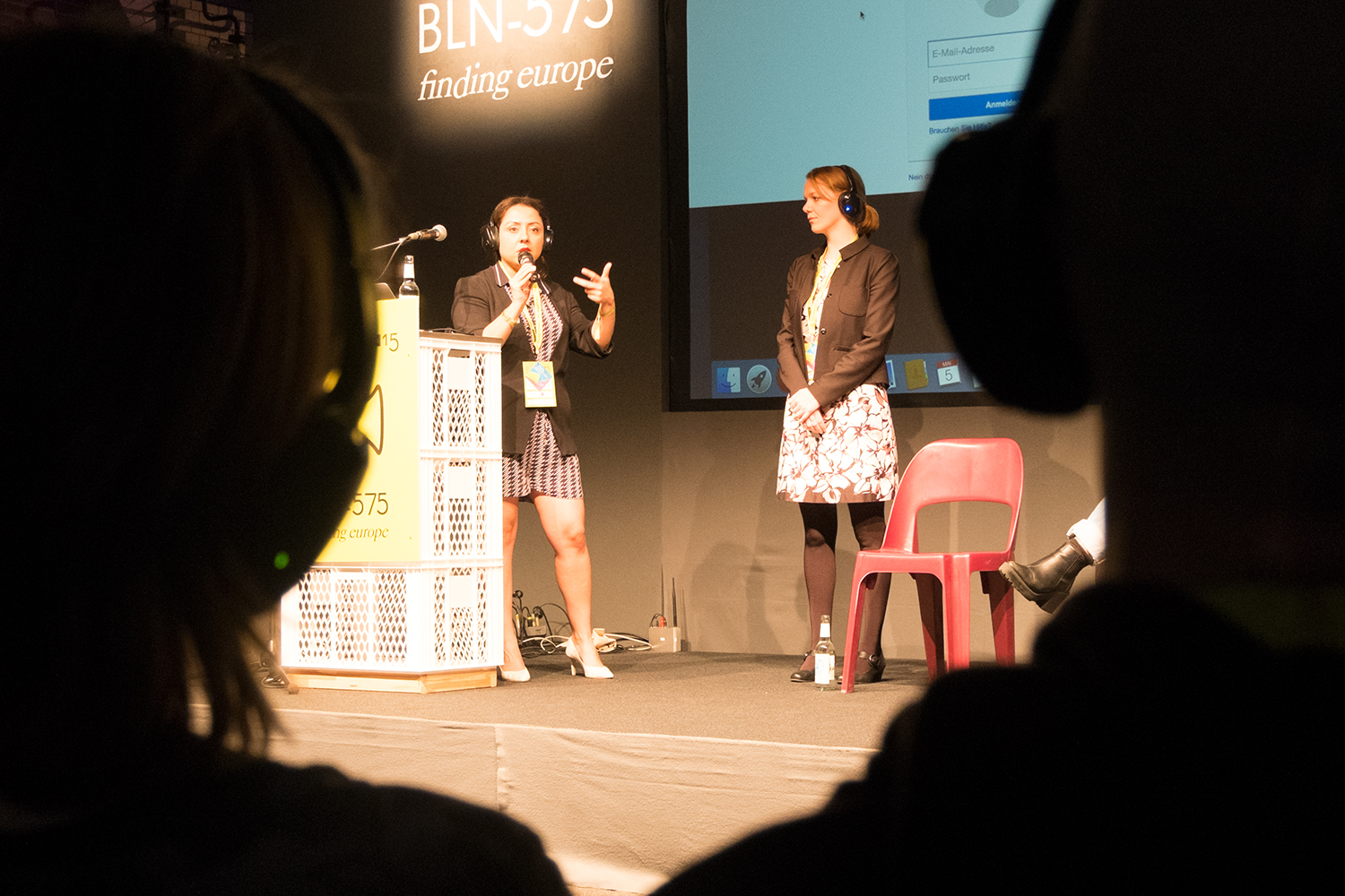 Photo of two women speaking on stage from behind the heads of two audience members