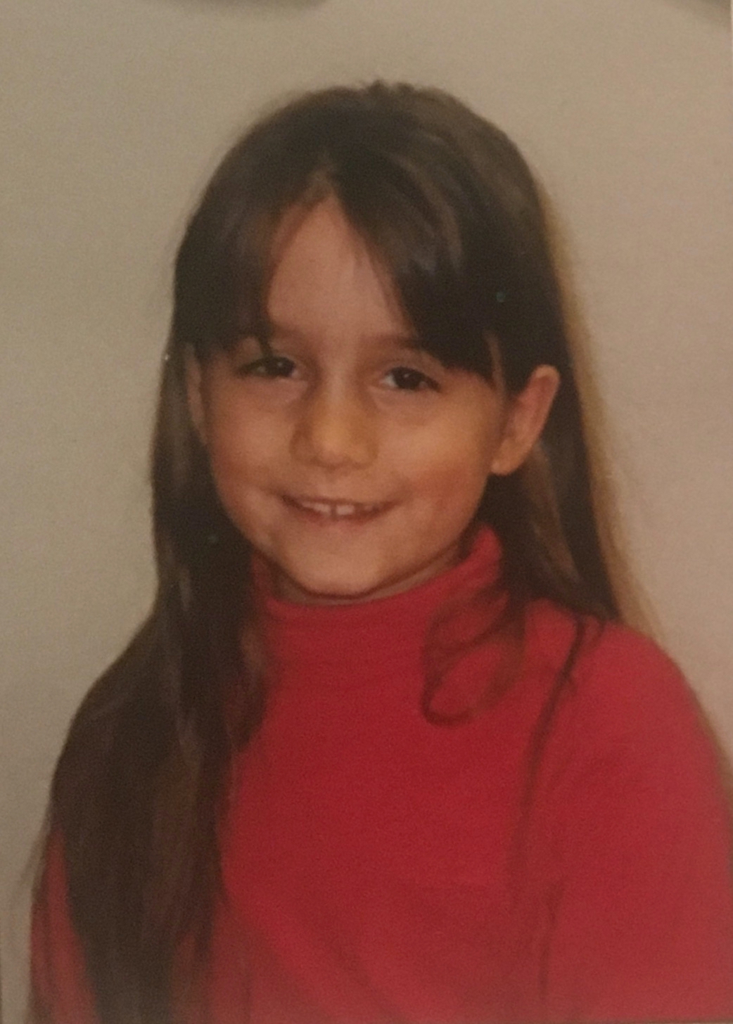 Portrait photo of me aged six, smiling with a long 90s fringe.